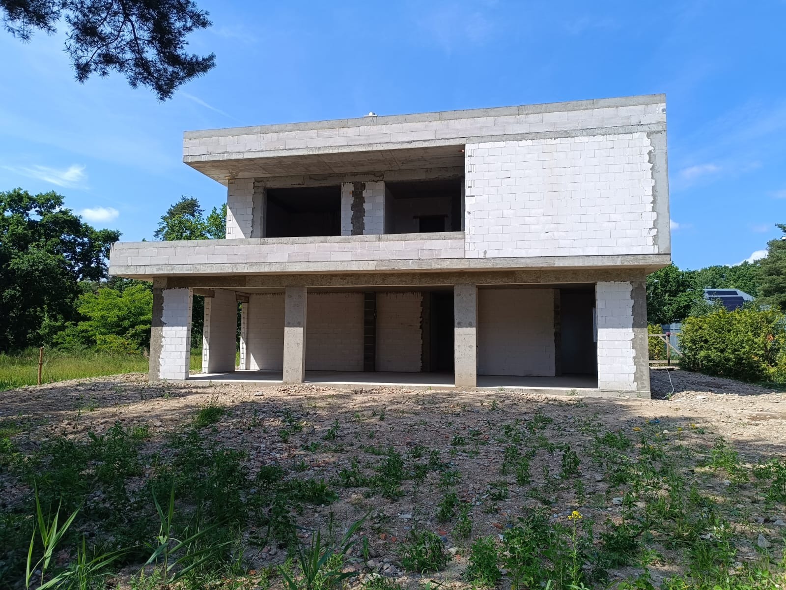 Dwukondygnacyjny dom w stanie surowym otwartym z białego bloczka, widok z zewnątrz na tle zieleni i błękitnego nieba, widać zarys przyszłych okien i drzwi.