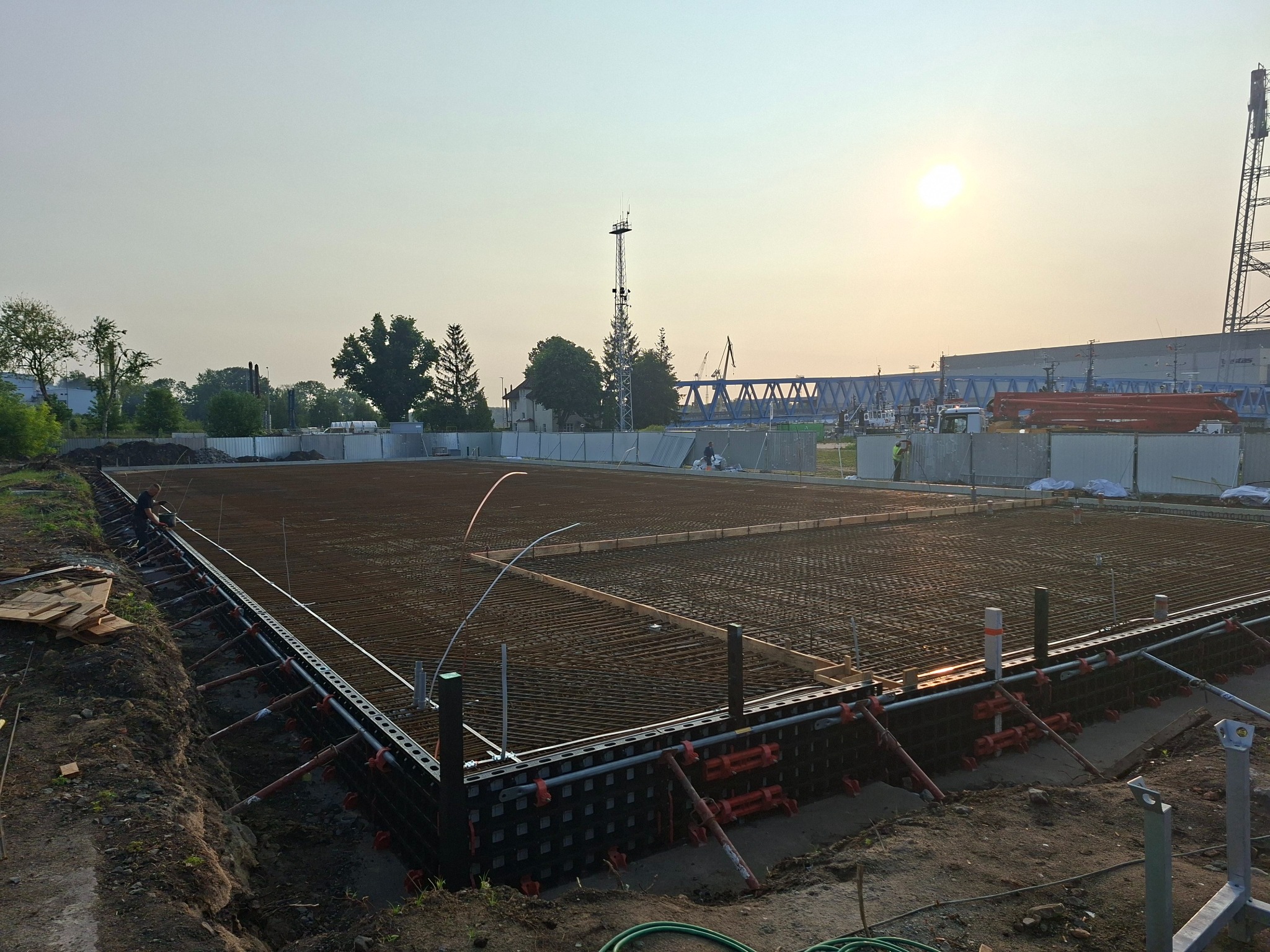 Zbrojenie fundamentów pod budowę, widoczne szalunki i siatka z prętów stalowych na tle zachodzącego słońca i przemysłowego krajobrazu.