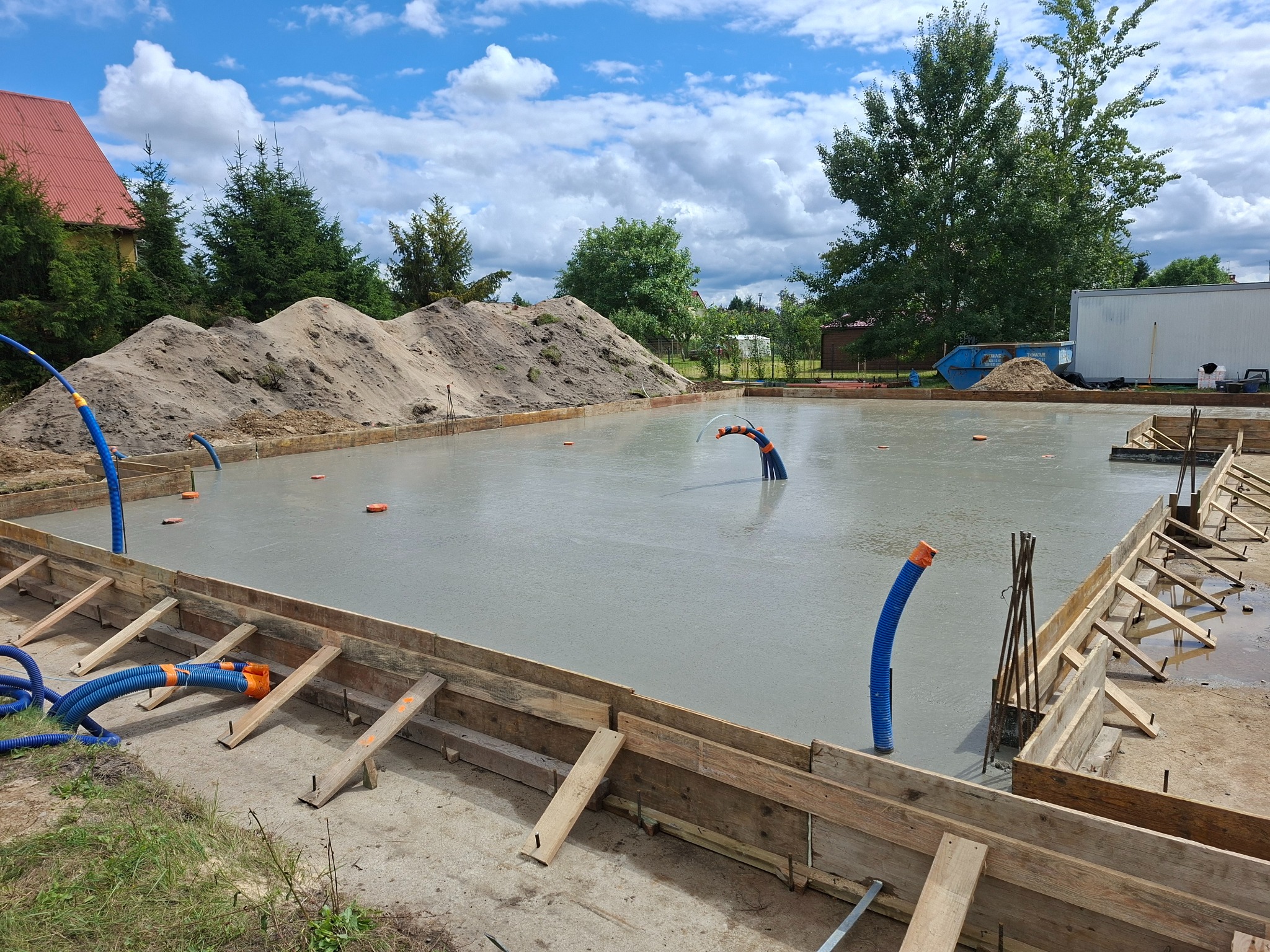 Zalany fundament z drewnianym szalunkiem, wystające rury instalacyjne. W tle hałda piasku i budynek z czerwonym dachem pod błękitnym niebem.
