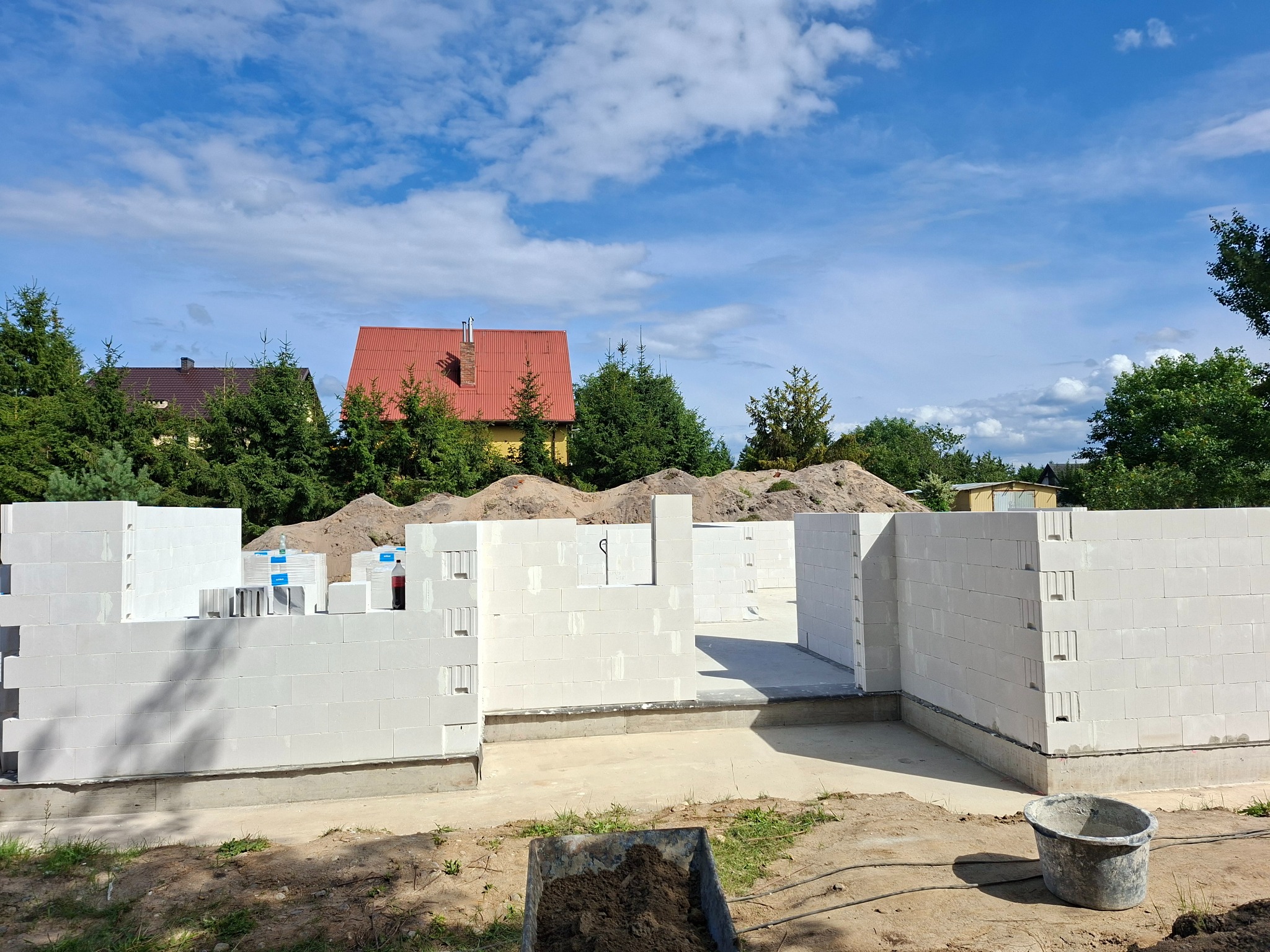 Wznoszone ściany parteru domu z białych bloczków, widoczne narzędzia budowlane, w tle dom z czerwonym dachem i zielone drzewa pod błękitnym niebem.