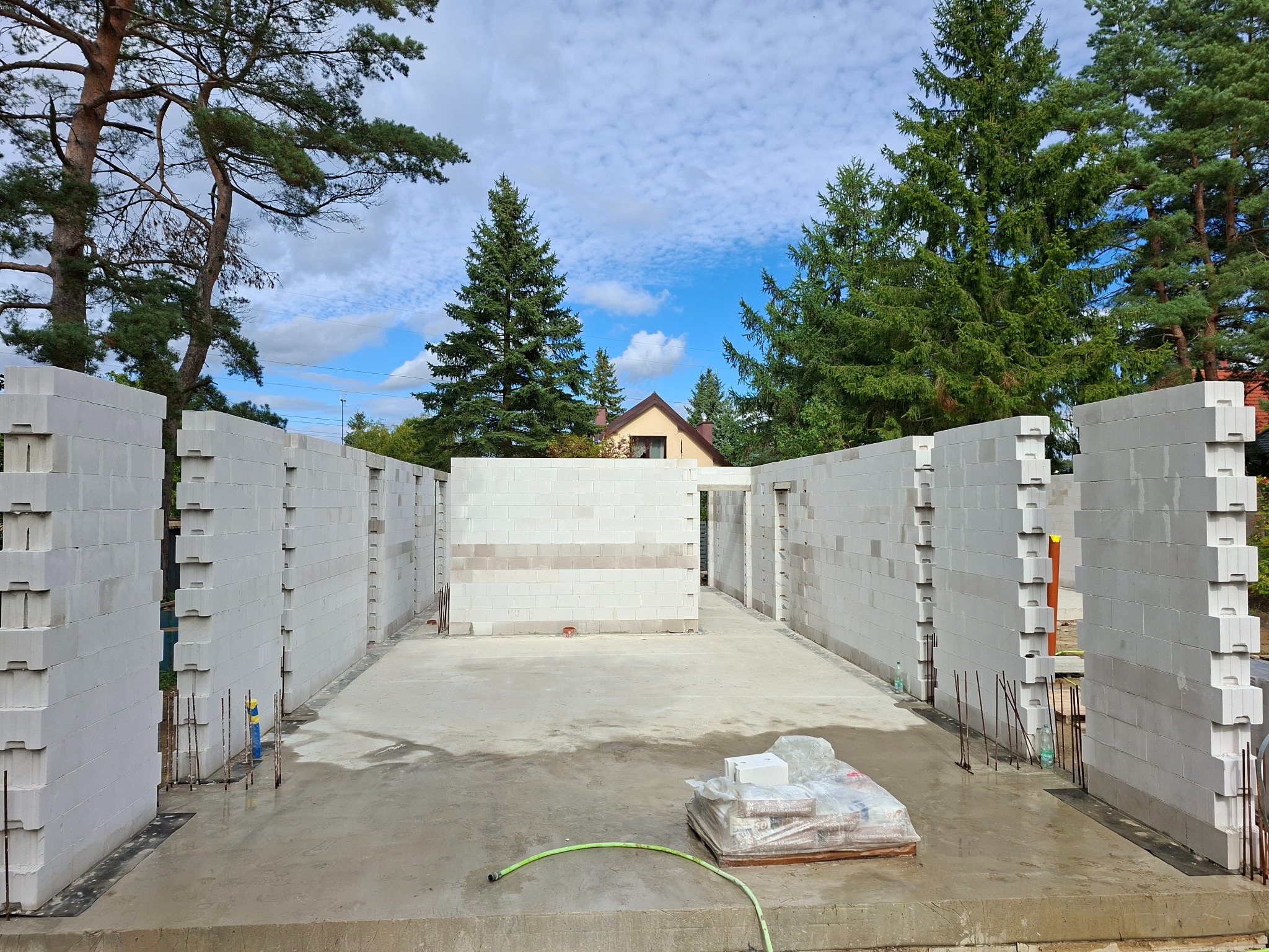 Wznoszenie ścian parteru z betonu komórkowego Ytong w Szczecinie. Widoczne spoiny pionowe i poziome, zbrojenie w otworach. W tle zieleń drzew i fragment dachu.