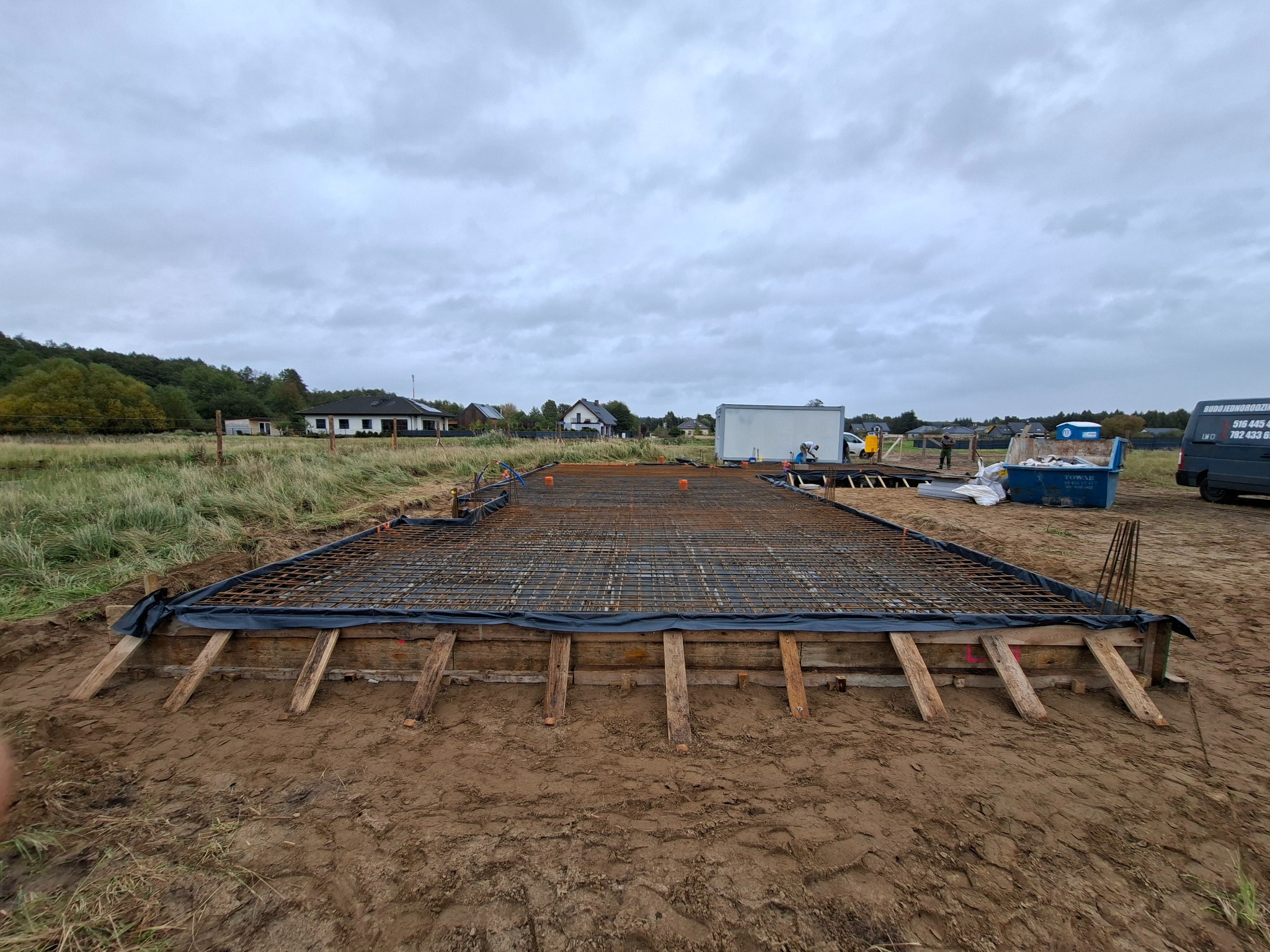 Zbrojenie fundamentu pod budowę domu jednorodzinnego, widok na siatkę zbrojeniową w drewnianym szalunku, przygotowaną do zalania betonem na tle pochmurnego nieba.