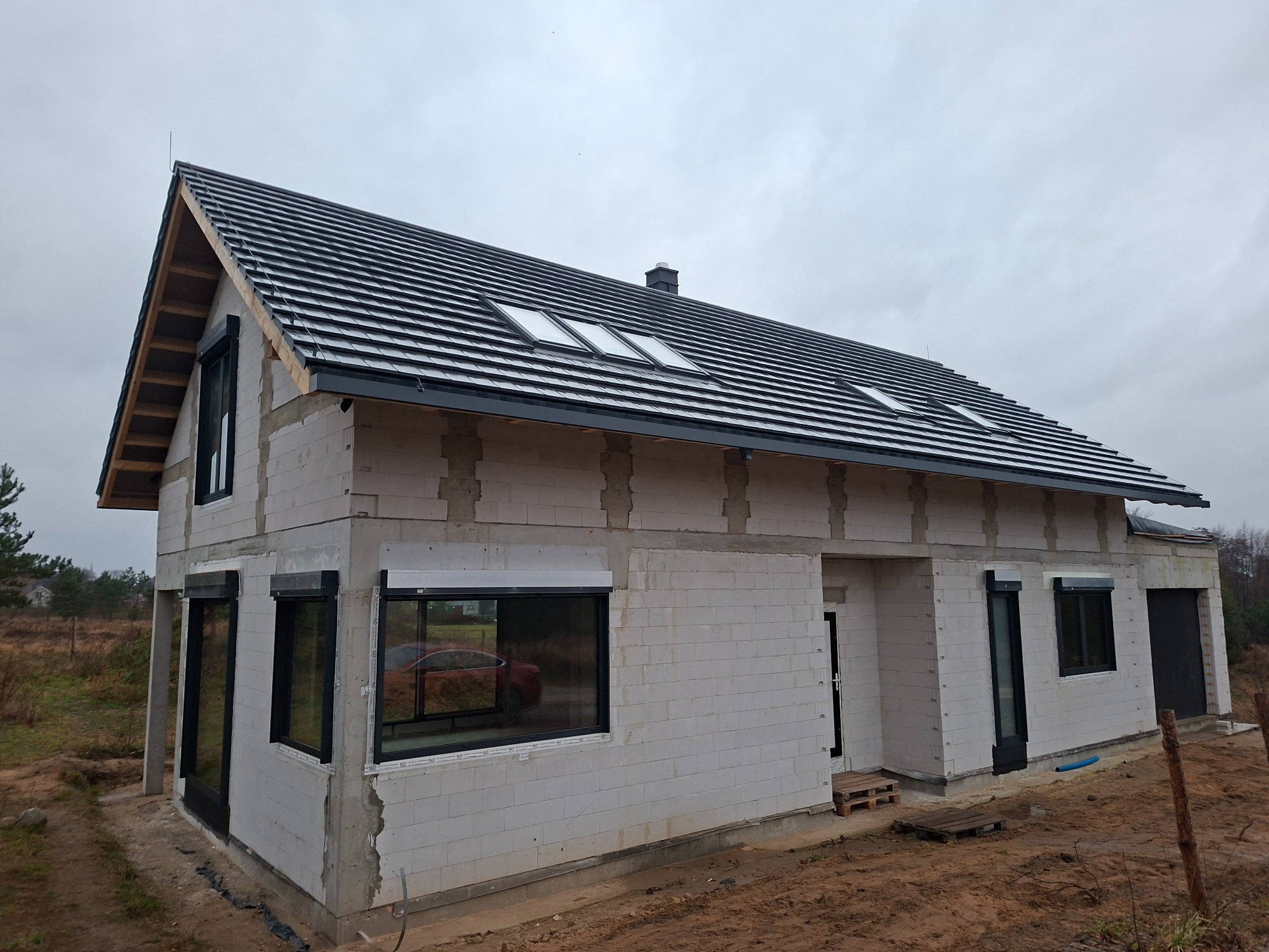 Niedokończony dom z białych bloczków, czarną dachówką i oknami z roletami zewnętrznymi. Widoczne elementy konstrukcyjne i brak elewacji. Szary dzień.