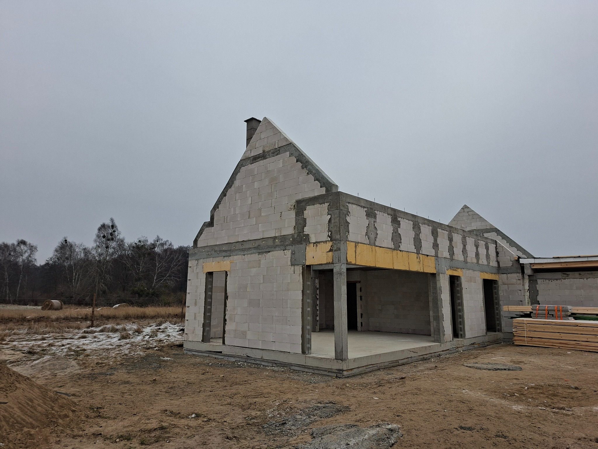 Surowy stan domu z białych bloczków, widoczne otwory okienne i drzwiowe, betonowe nadproża, w tle drzewa i częściowo zaśnieżone pole. Budowa w toku.