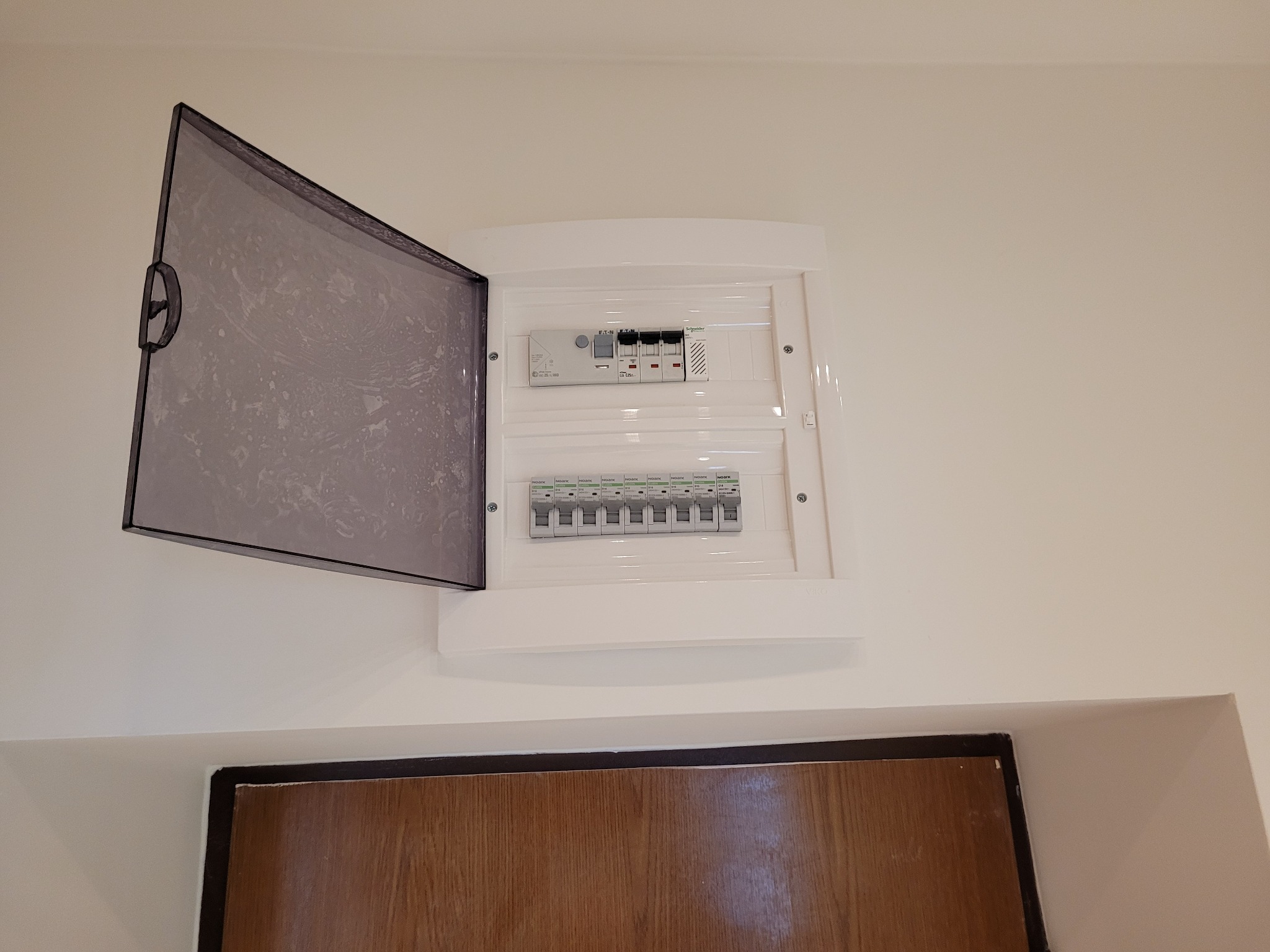 Rozdzielnica elektryczna natynkowa z otwartą, przydymioną pokrywą, widoczne bezpieczniki i moduły, zamontowana na białej ścianie nad futryną drzwi.