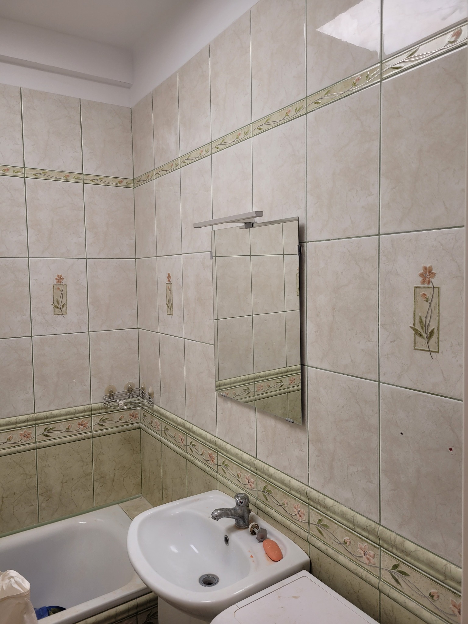 Ukończona łazienka z beżowymi płytkami z motywem kwiatowym, widoczne umywalka, wanna i lustro z oświetleniem, brak baterii przy wannie, mydło na umywalce.