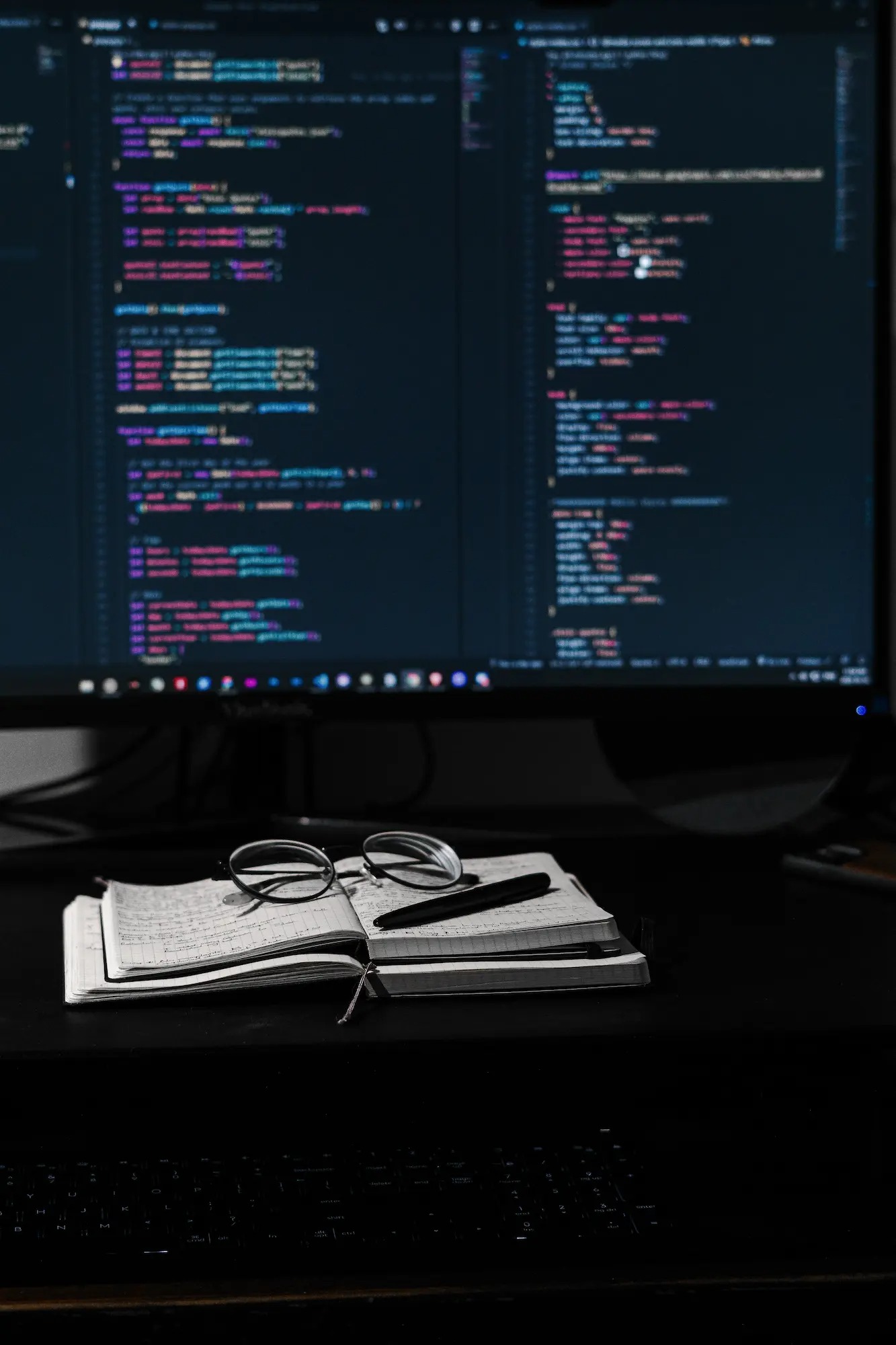 Monitor wyświetlający kod programistyczny z notatnikiem, okularami i długopisem na biurku w słabym oświetleniu.