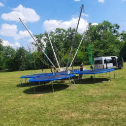 Rozstawiony park trampolin typu bungee na trawie w słoneczny dzień, biały bus zaparkowany obok.