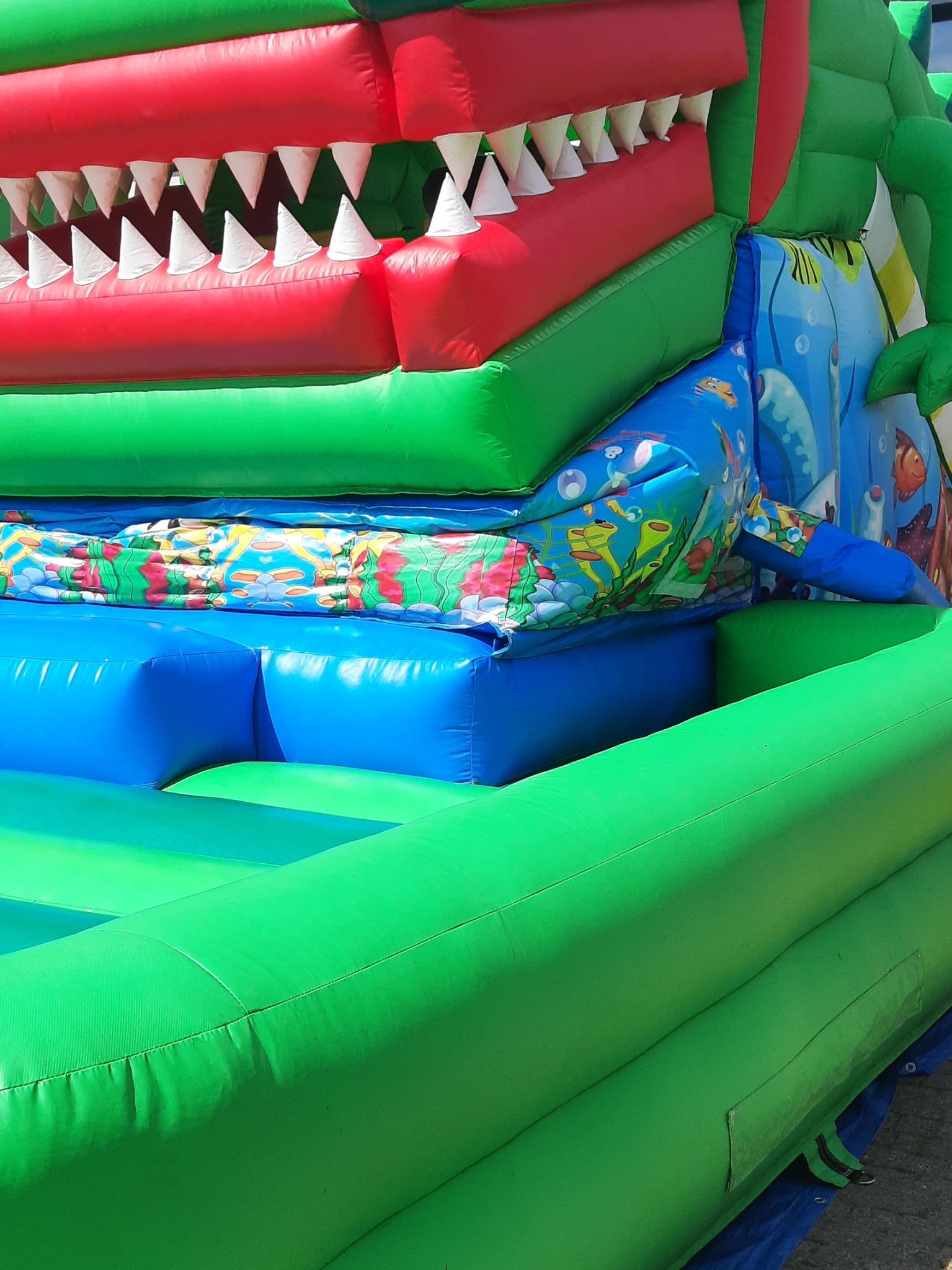 Fragment dmuchanego zamku w kształcie paszczy krokodyla z widocznymi, trójkątnymi zębami, zielonymi bokami i niebieskim wnętrzem z motywem podwodnego świata.