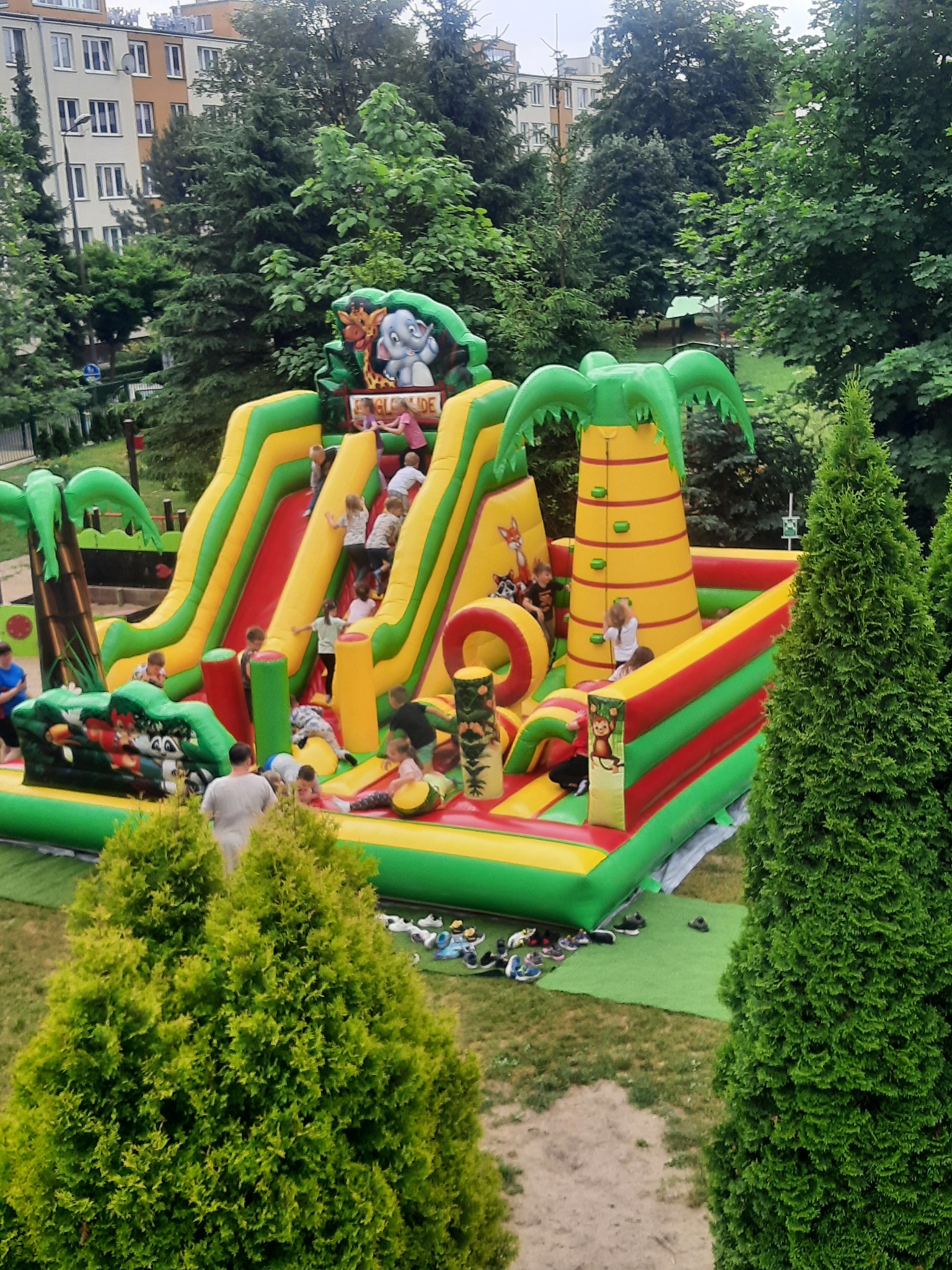 Dzieci bawią się na dmuchanym placu zabaw w motywie dżungli z palmami i zjeżdżalniami na tle bloków mieszkalnych i drzew.