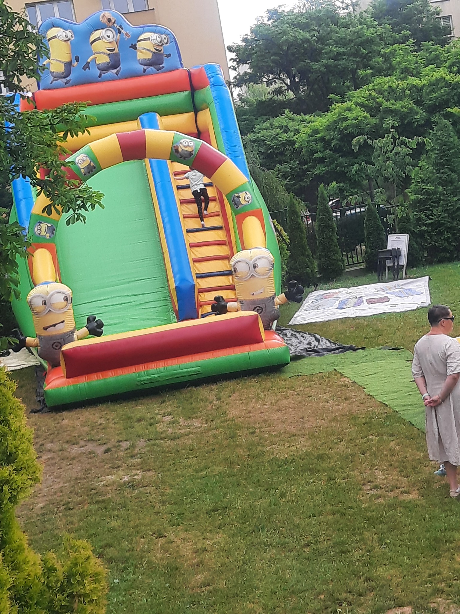 Dziecko wchodzi po schodach na dmuchaną zjeżdżalnię z motywem Minionków, ustawioną na trawniku w ogrodzie.
