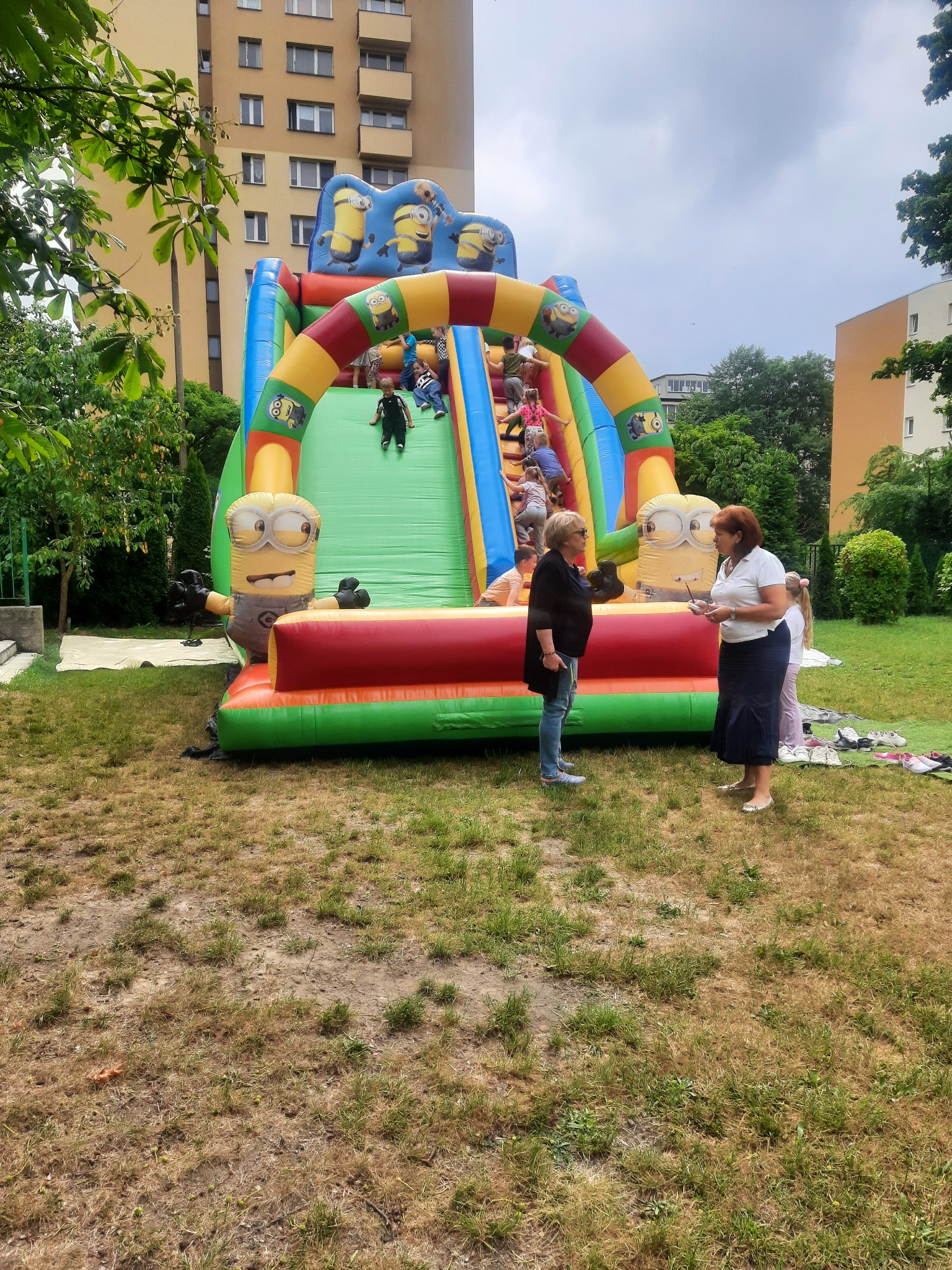 Dmuchany zamek ze zjeżdżalnią w motywie Minionków, z dziećmi bawiącymi się na nim, oraz dwiema kobietami rozmawiającymi w pobliżu na trawniku.