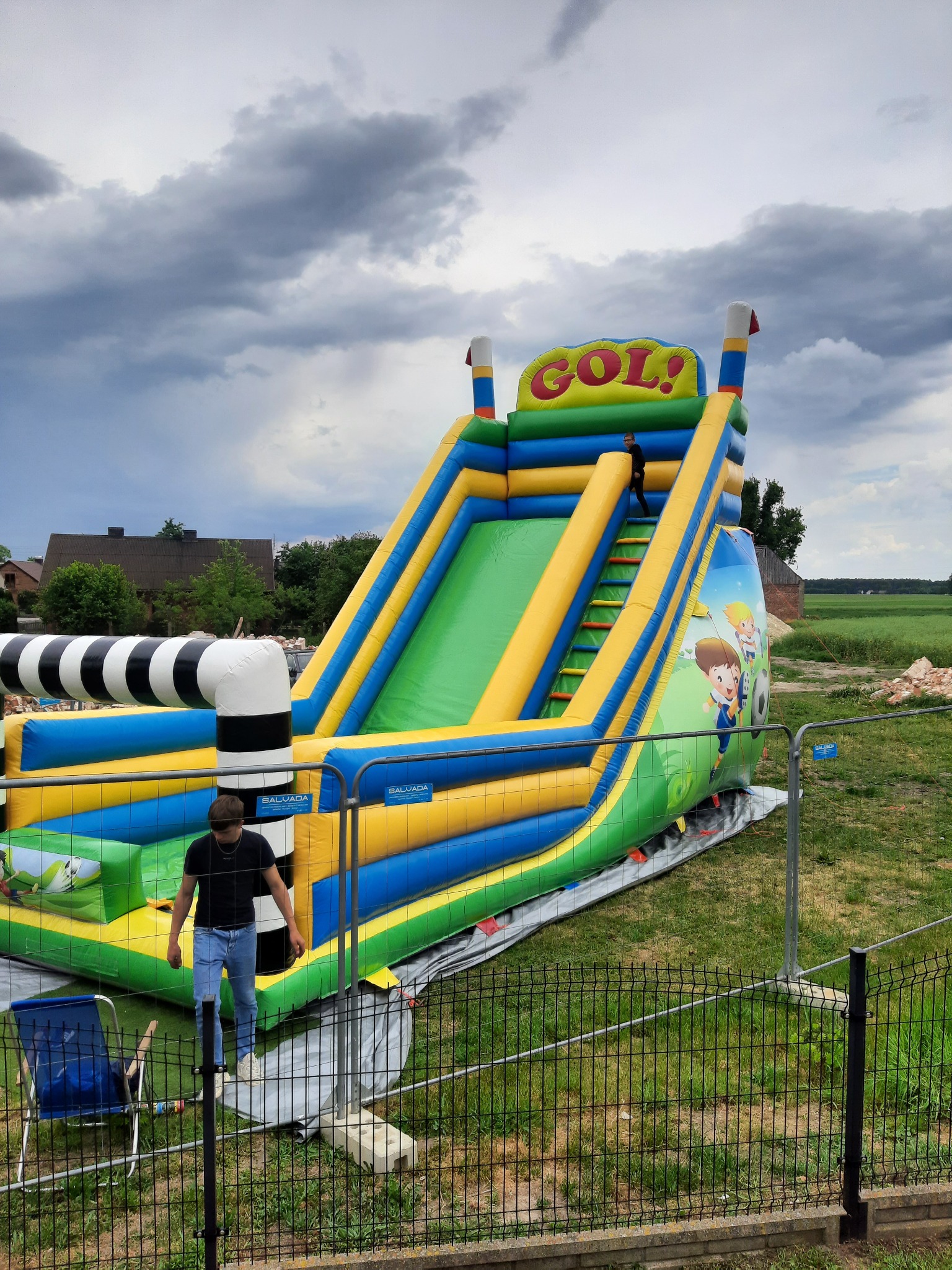 Duża, kolorowa dmuchana zjeżdżalnia z napisem 'GOL!' na tle pochmurnego nieba, z dwójką dzieci i ogrodzeniem.