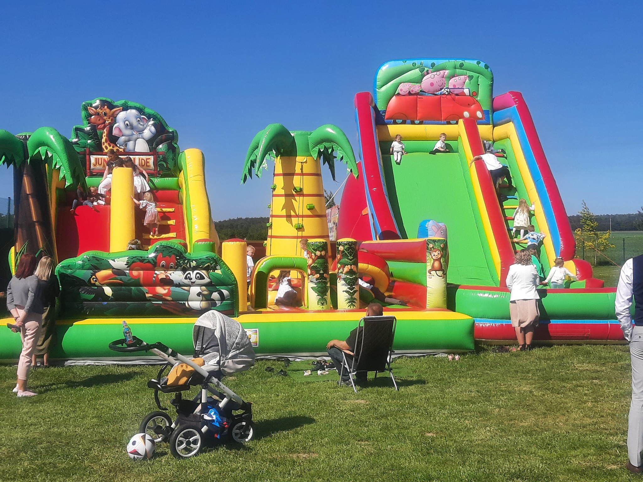 Dwa kolorowe, dmuchane zamki z motywami zwierzęcymi i postaci z bajek, dzieci bawiące się na zamkach, wózek dziecięcy i osoby dorosłe na trawniku w słoneczny dzień.