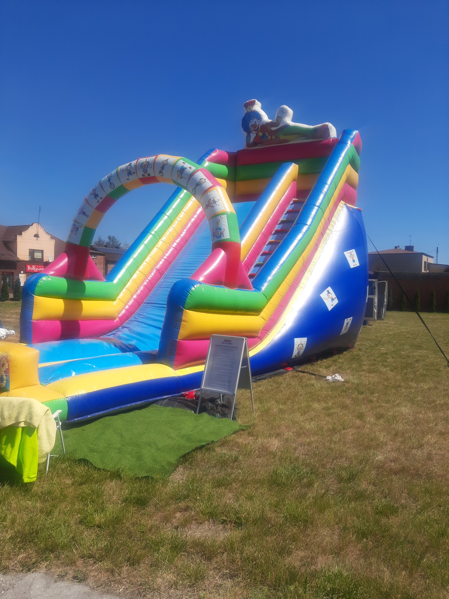 Kolorowa, dmuchana zjeżdżalnia z motywem klauna na szczycie, ustawiona na trawniku w słoneczny dzień. Widoczny baner informacyjny obok i częściowo zabudowania w tle.