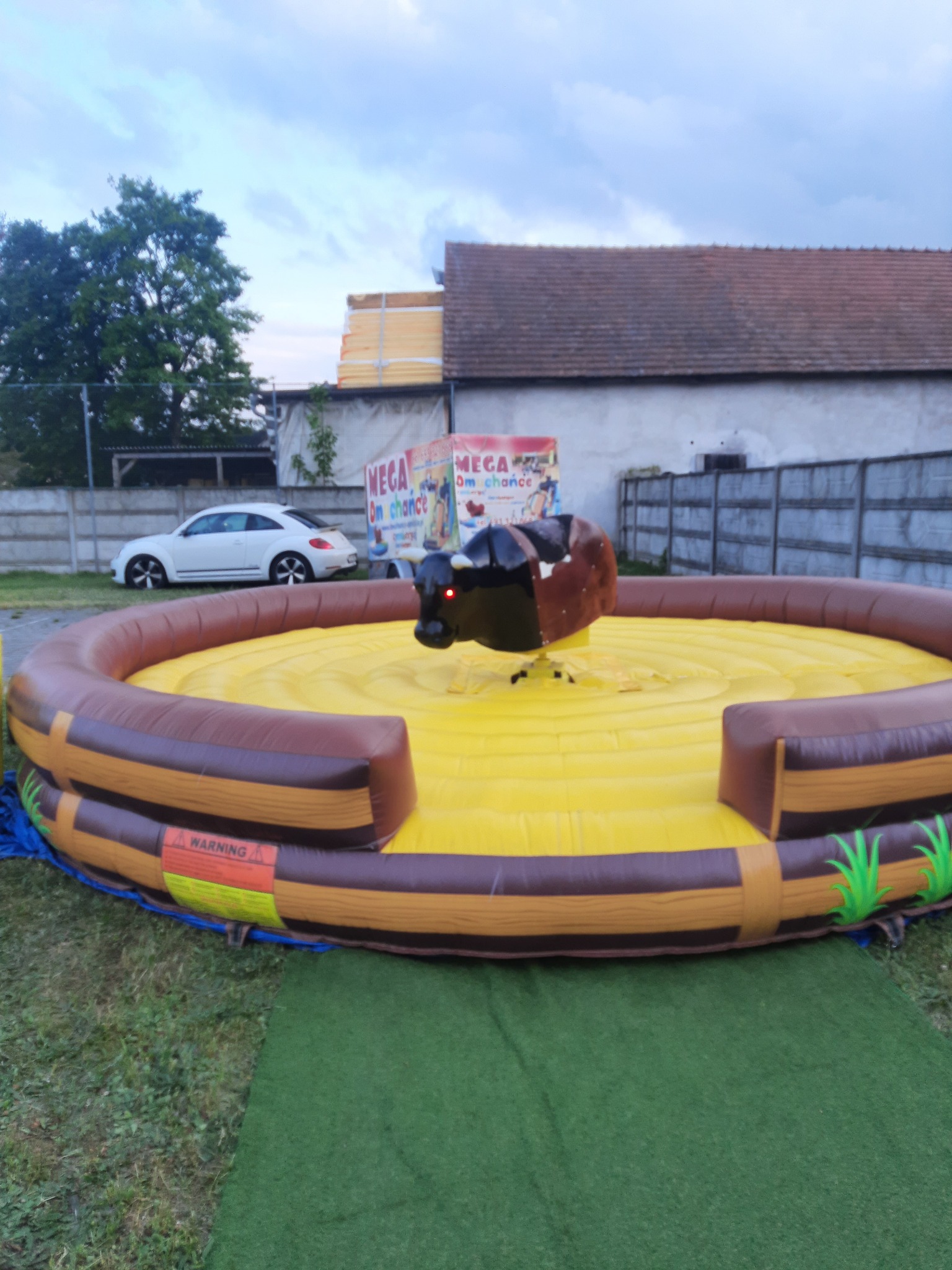 Mechaniczny byk na dmuchanym, żółtym ringu z brązowymi elementami imitującymi drewno, ustawiony na trawie, z białym samochodem i budynkami w tle.