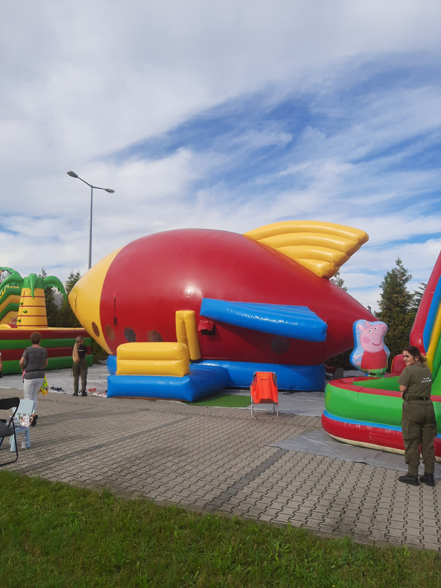 Duży, nadmuchiwany statek kosmiczny w kolorach czerwieni i żółci, z elementami niebieskimi i postacią świnki Peppy, ustawiony na utwardzonym placu podczas imprezy plenerowej, w tle inne dmuchańce...