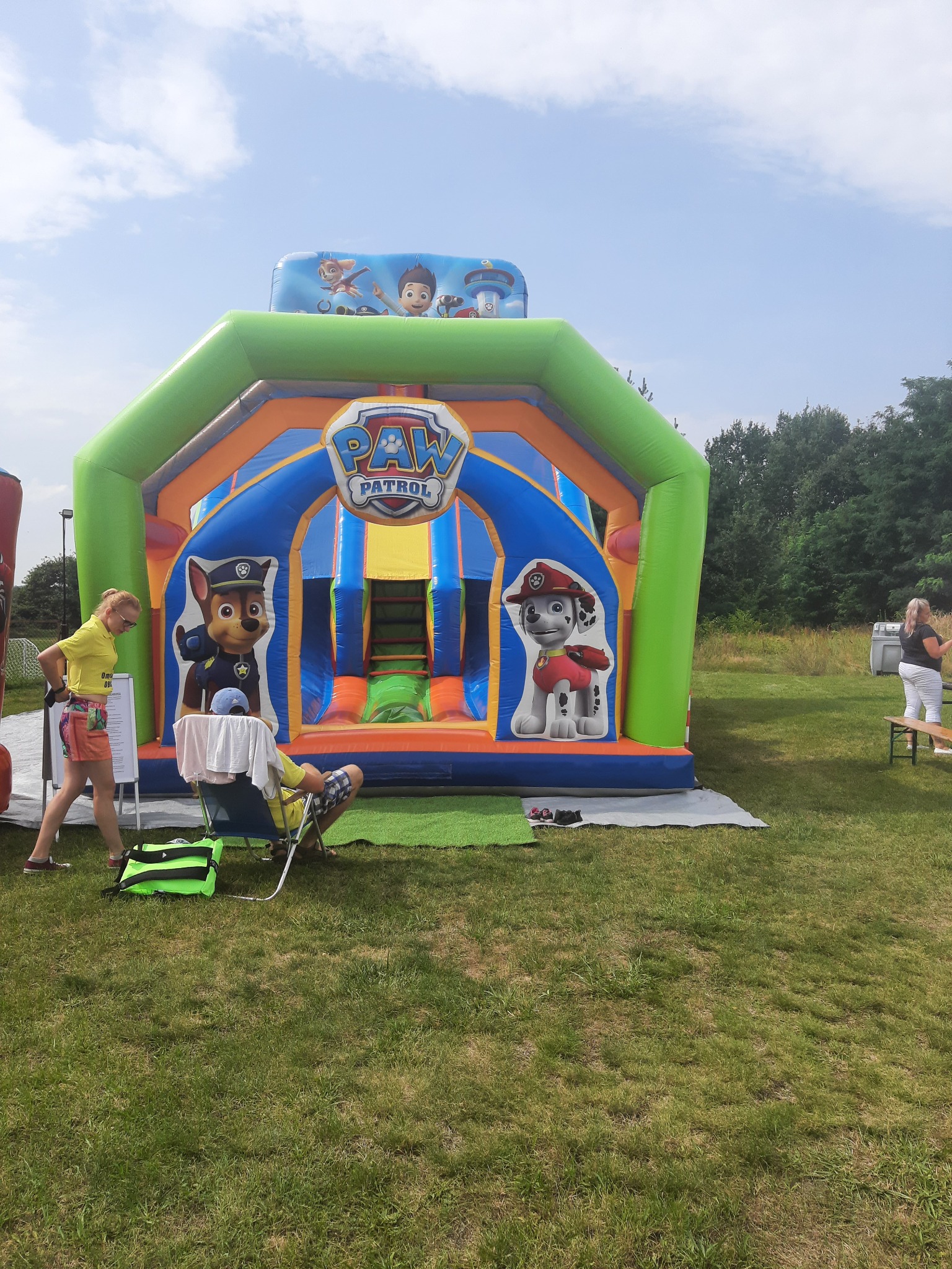 Kolorowy dmuchany zamek z motywem 'Psi Patrol' na trawniku, obok stoi kobieta z listą i mężczyzna siedzący na krześle.