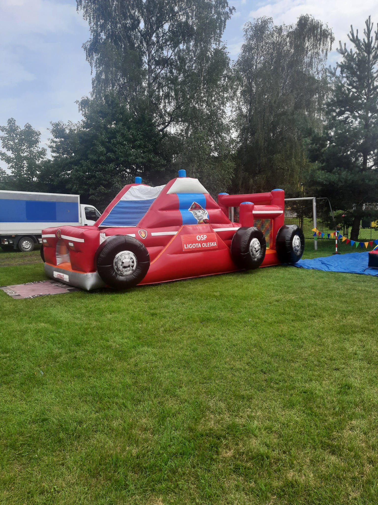 Czerwony dmuchany zamek w kształcie wozu strażackiego z napisem 'OSP Ligota Oleska' na trawie, z widocznym fragmentem boiska i samochodu w tle.