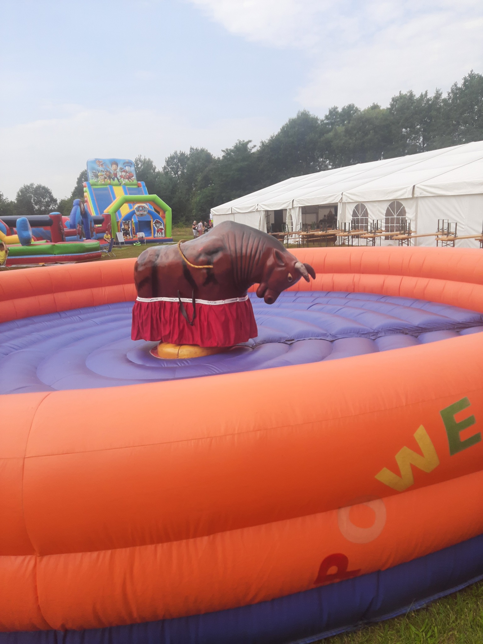 Mechaniczny byk na dmuchanym pomarańczowo-niebieskim podeście z napisem 'POWER', w tle dmuchany plac zabaw dla dzieci i biały namiot.