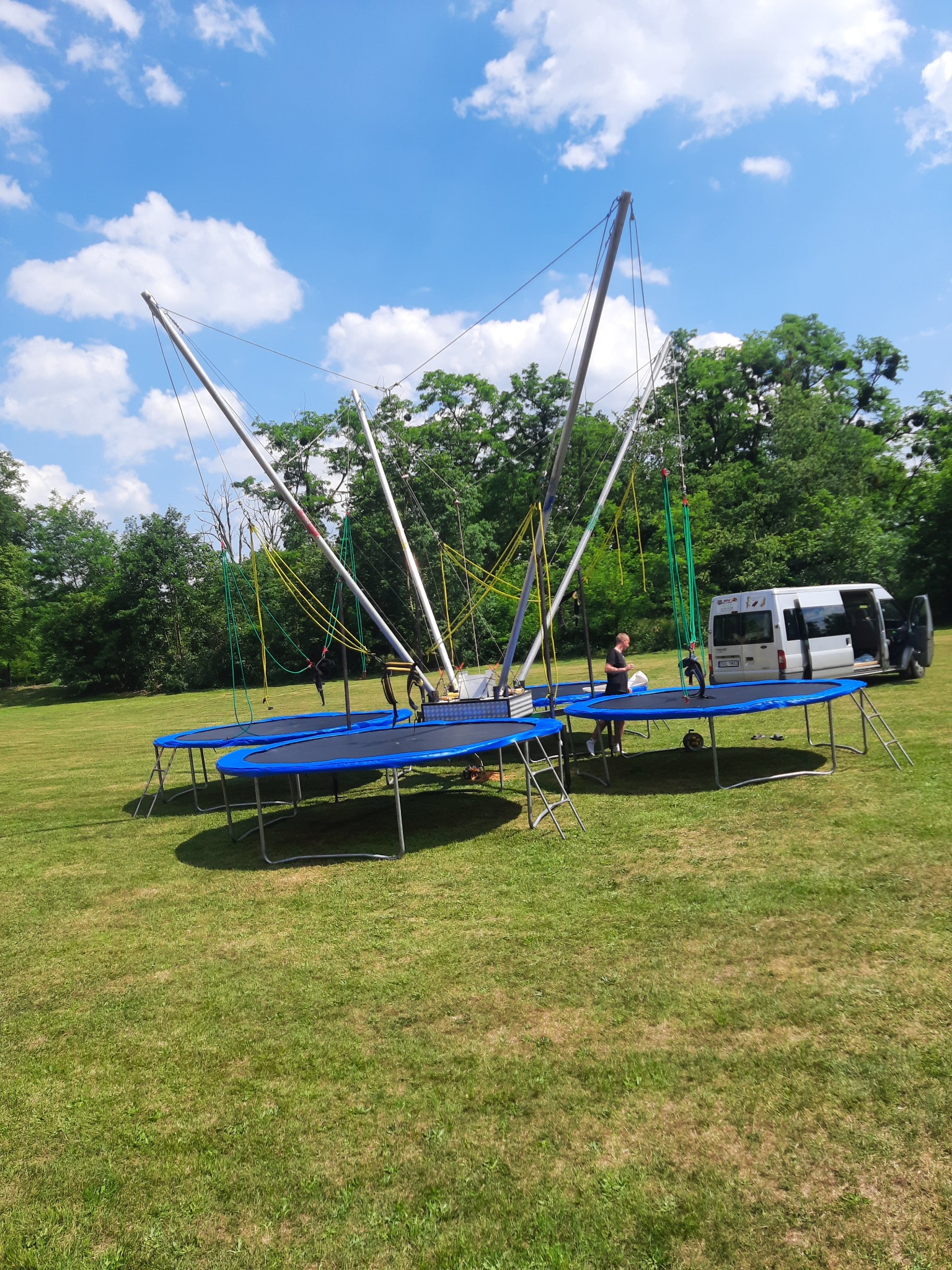 Rozstawiony park trampolin typu bungee na trawie w słoneczny dzień, biały bus zaparkowany obok.