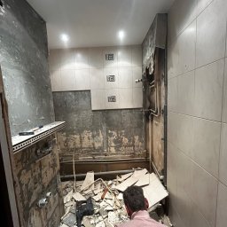 LILAY MONIKA JAKÓBCZYK - Demontaż starej glazury w łazience, widoczne fragmenty płytek na podłodze i ścianach, odsłonięte rury, mężczyzna w różowej bluzie z tyłu.