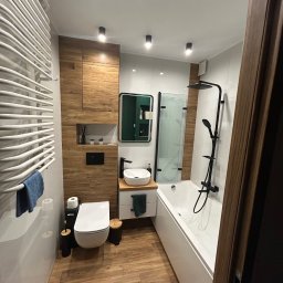 LILAY MONIKA JAKÓBCZYK - Nowoczesna łazienka z drewnopodobnymi płytkami, białą armaturą i czarnymi akcentami, w tym bateria umywalkowa i deszczownica z natryskiem.