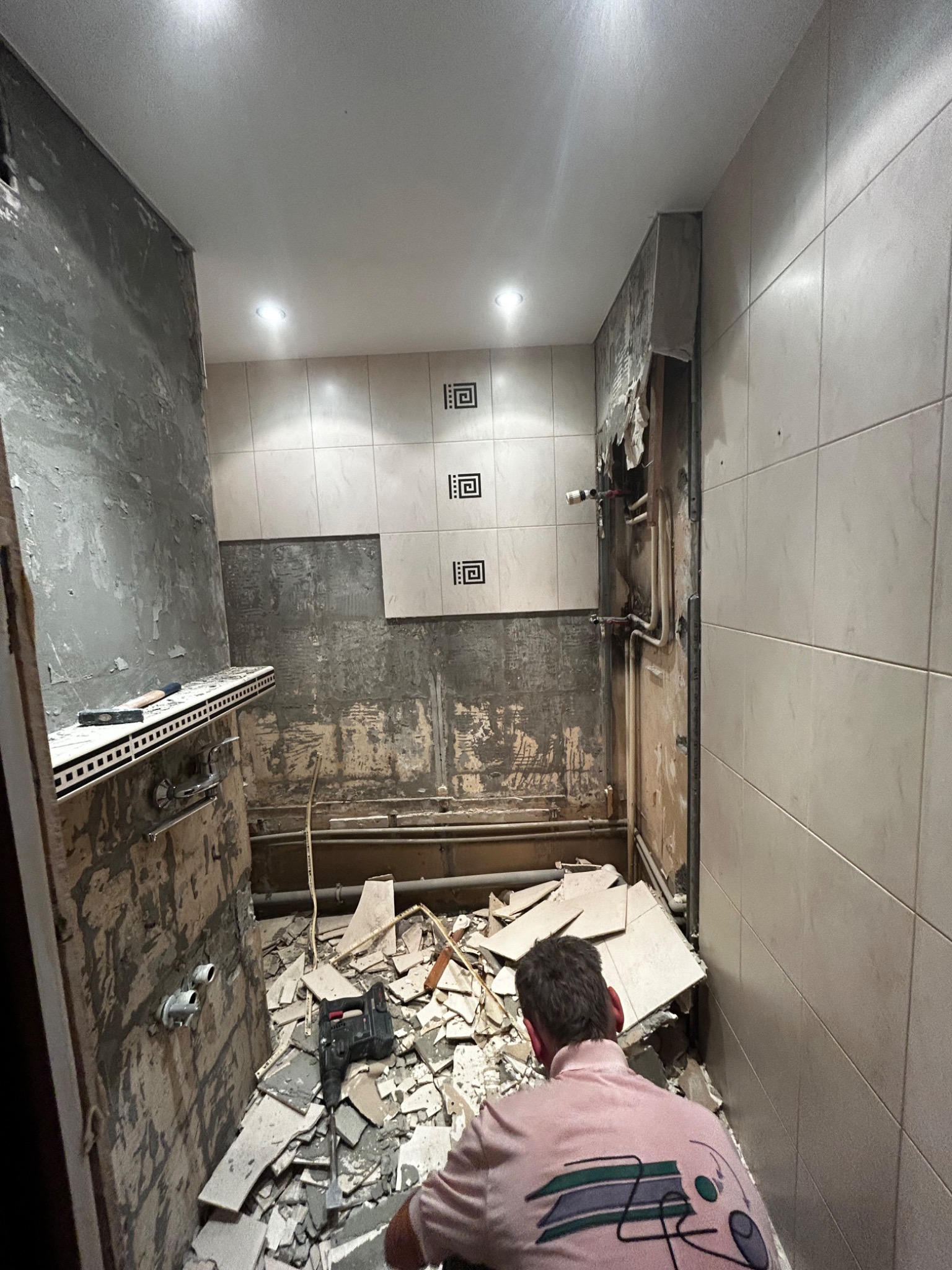 Demontaż starej glazury w łazience, widoczne fragmenty płytek na podłodze i ścianach, odsłonięte rury, mężczyzna w różowej bluzie z tyłu.