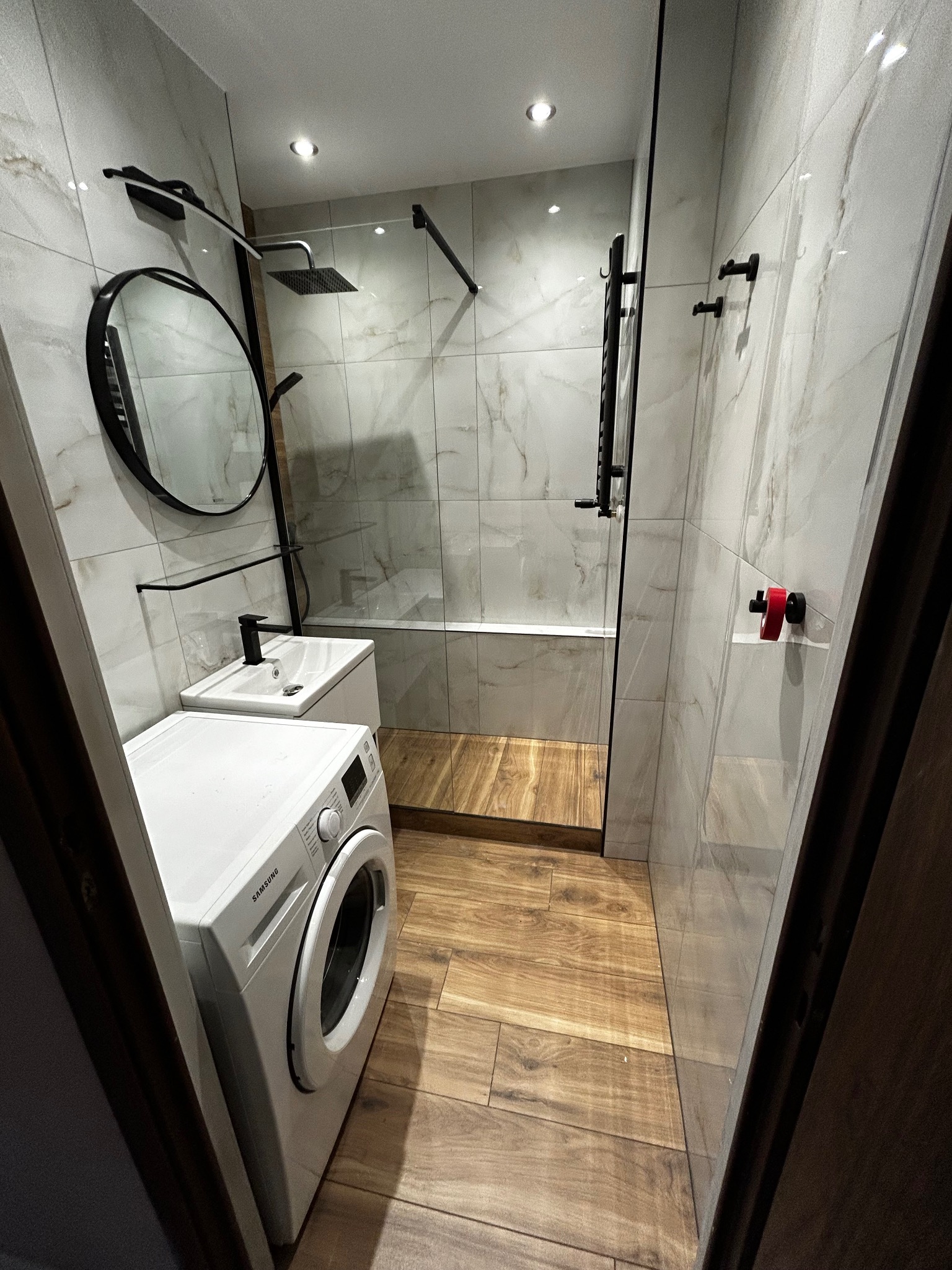 Nowoczesna łazienka z kabiną prysznicową typu walk-in, wykończona płytkami imitującymi marmur, drewnopodobną podłogą i czarnymi akcentami armatury, z pralką Samsung w rogu.