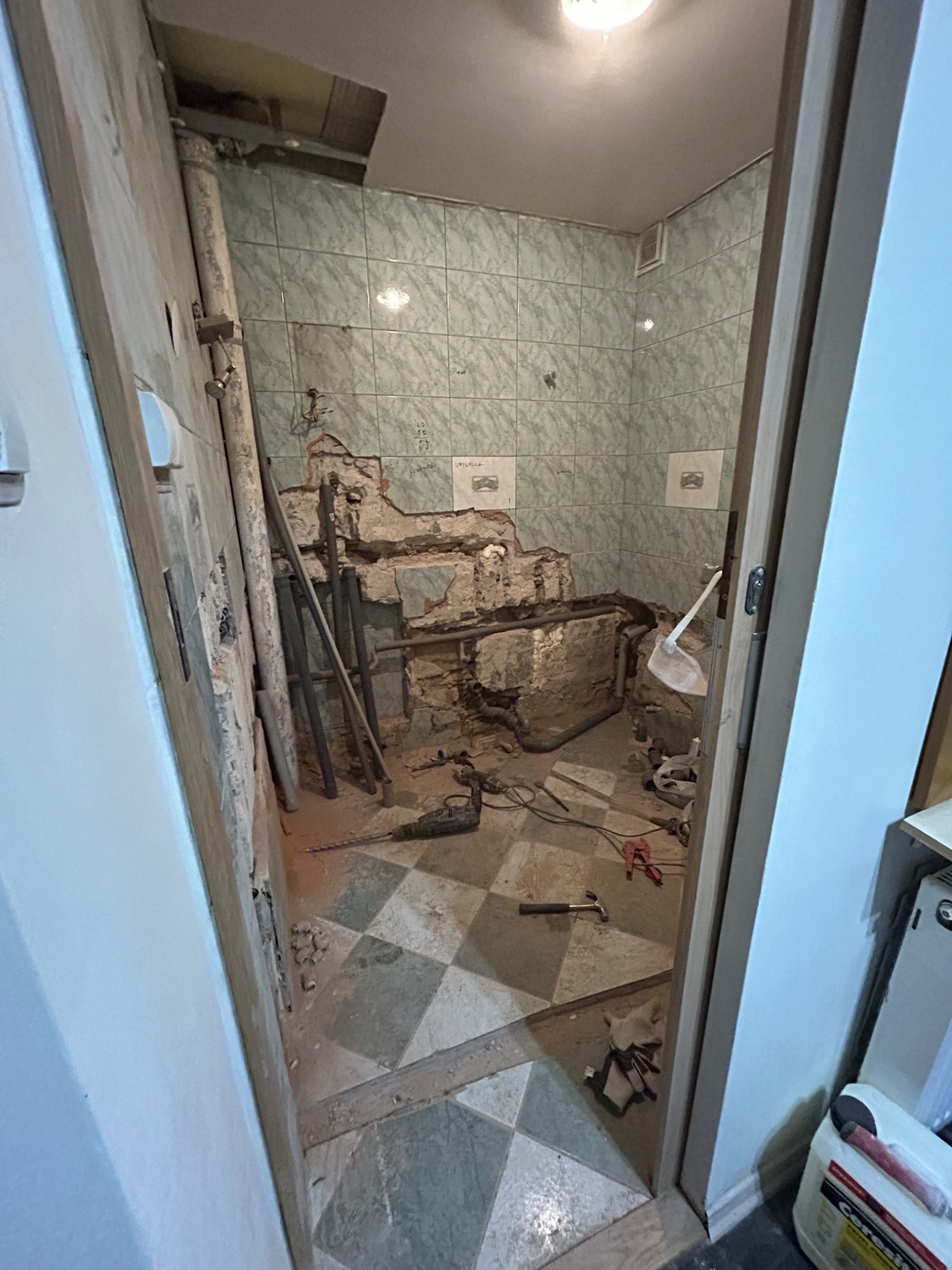 Łazienka w trakcie demontażu: zerwane płytki, odsłonięte rury i instalacje, widoczny gruz i narzędzia na podłodze w szachownicę, częściowo zdemontowana ściana z widocznymi napisami.