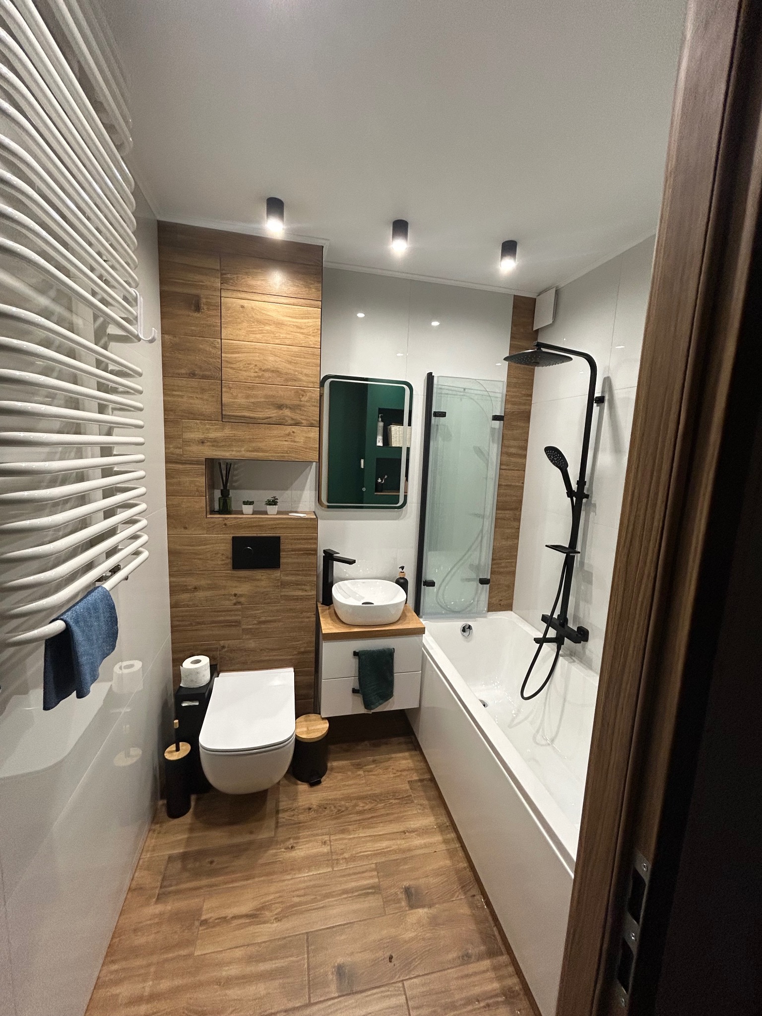 Nowoczesna łazienka z drewnopodobnymi płytkami, białą armaturą i czarnymi akcentami, w tym bateria umywalkowa i deszczownica z natryskiem.