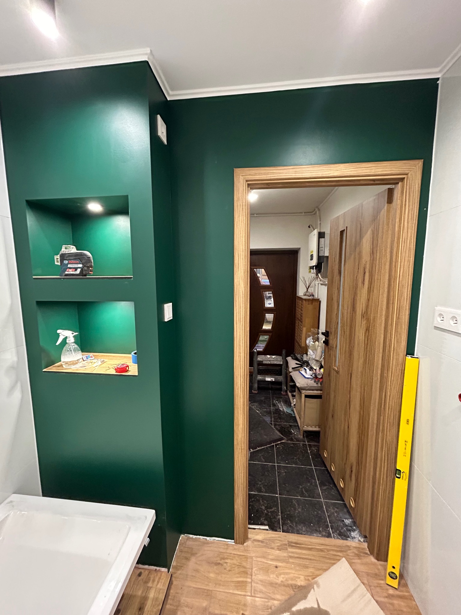 Zielona ściana z wnękami oświetlonymi LED, w jednej poziomica laserowa Bosch, w drugiej butelka ze spryskiwaczem i taśma miernicza, widoczny fragment brodzika i drzwi wewnętrzne z ościeżnicą...