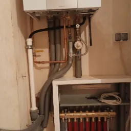 Wnętrze kotłowni z wiszącym, nowoczesnym kotłem gazowym marki ecoTEC plus, widoczne rury miedziane i szare izolowane, oraz rozdzielacz ogrzewania podłogowego z czerwonymi i niebieskimi pętlami.