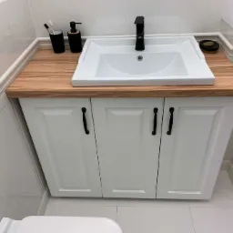 Biała szafka łazienkowa pod umywalkę z drewnianym blatem w odcieniu ciepłego drewna, czarna bateria i akcesoria łazienkowe, widok z perspektywy podłogi.