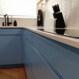 Nowoczesna kuchnia z błękitnymi szafkami, drewnianą podłogą w jodełkę i białymi płytkami ułożonymi w chevron. Na blacie stoją czarne akcesoria kuchenne, oświetlenie LED pod górnymi szafkami.