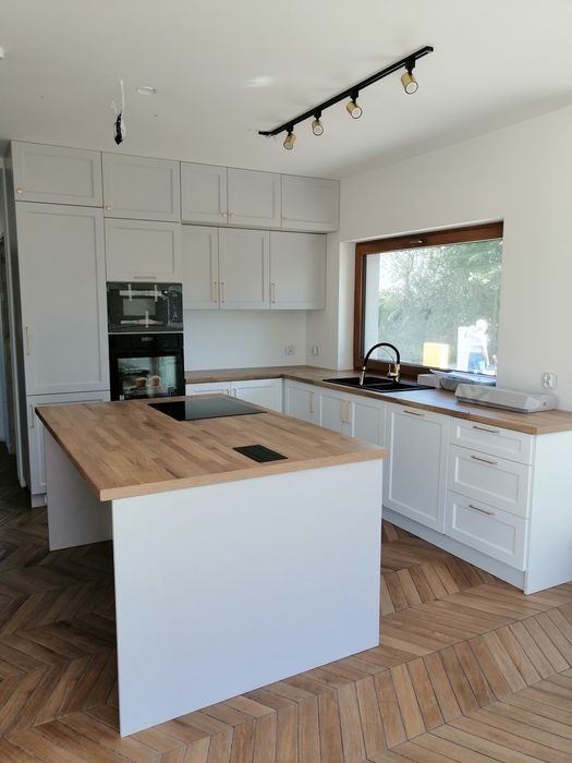 Jasna kuchnia z wyspą w Elblągu, drewniany blat i parkiet w jodełkę, nowoczesne oświetlenie punktowe.