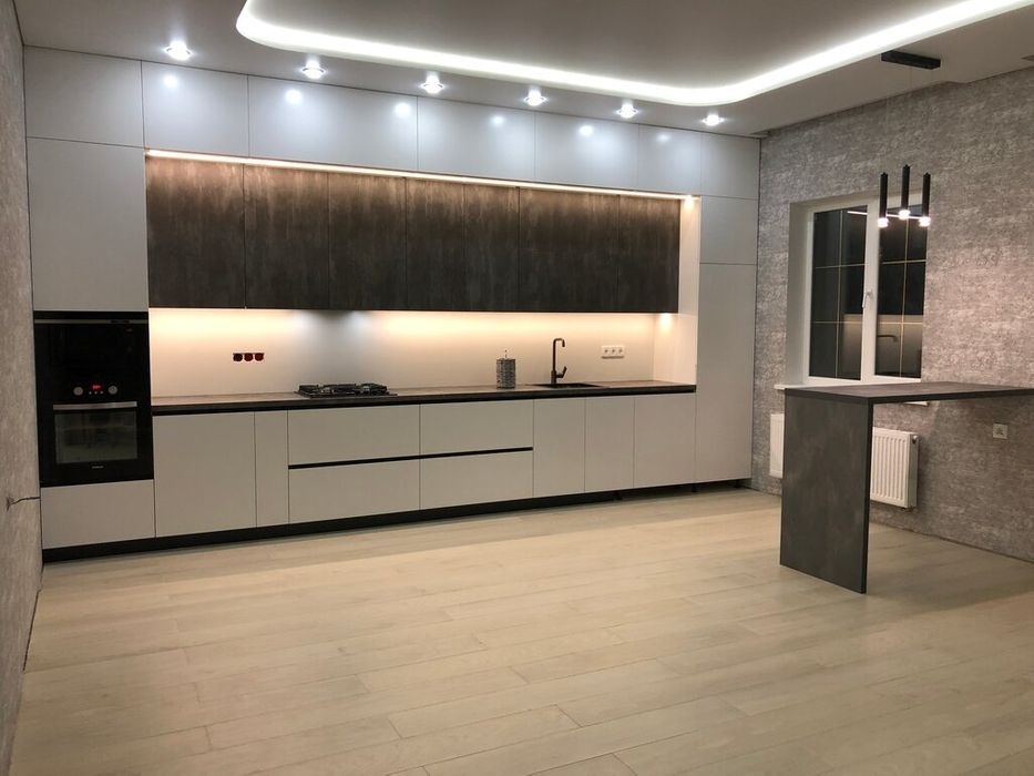 Nowoczesna kuchnia z białymi szafkami dolnymi i ciemnoszarymi górnymi, podświetlonymi listwami LED, z czarną armaturą i blatem, uzupełniona o wysoki stół barowy w tym samym odcieniu szarości.