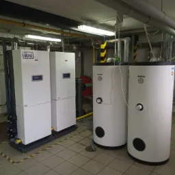 Dwa białe kotły grzewcze marki Tilgner z wyświetlaczami i rurami, oraz dwa białe zasobniki ciepłej wody Kospel z manometrami, zainstalowane w kotłowni z kafelkową podłogą i rurami pod sufitem.