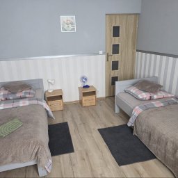 Centrum Szkoleniowo-Terapeutyczne Life - Dwa szare łóżka z brązowymi narzutami i pościelą w kratę w pokoju z szaro-białą tapetą w pasy. Między nimi drewniane szafki nocne z lampką i wentylatorem, na ścianie obraz z tulipanami.