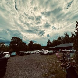 Centrum Szkoleniowo-Terapeutyczne Life - Szeroki kadr żwirowego parkingu z rzędem samochodów, obok budynek z ciemnym dachem i grupą ludzi. Na pierwszym planie różowy fotel i narzędzia. Nad całością dramatyczne, pofalowane chmury.