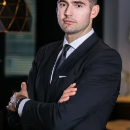 Portret pewnego mężczyzny w ciemnym garniturze, z rękami skrzyżowanymi na piersiach, w tle widoczna nowoczesna lampa.