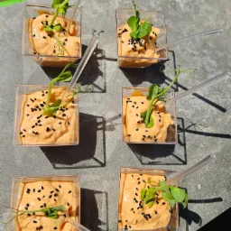 Sześć porcji kremowego musu w przezroczystych kwadratowych naczyniach, udekorowanych czarnym sezamem i młodymi pędami groszku, podane z przezroczystymi łyżeczkami.