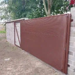 Przesuwna brama wjazdowa z poziomymi panelami w kolorze brązowym, zamontowana na murze z bloczków betonowych, widoczna z boku na tle zieleni.