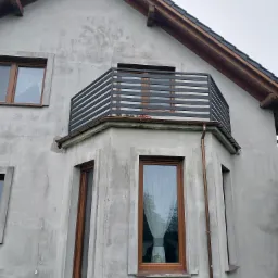 Szary dom jednorodzinny z balkonem z poziomymi, czarnymi balustradami, widok z dołu na elewację w trakcie wykańczania, widoczne okna z brązowymi ramami i rynna