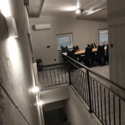 Wewnętrzna balustrada metalowa z czarnymi pionowymi prętami, oświetlenie ścienne LED, widok z góry schodów na jadalnię z dużym stołem i krzesłami.