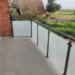 Balkon z nowoczesną balustradą szklaną z czarnymi słupkami, widok na wiejski krajobraz w tle, jasne płytki na podłodze balkonu.