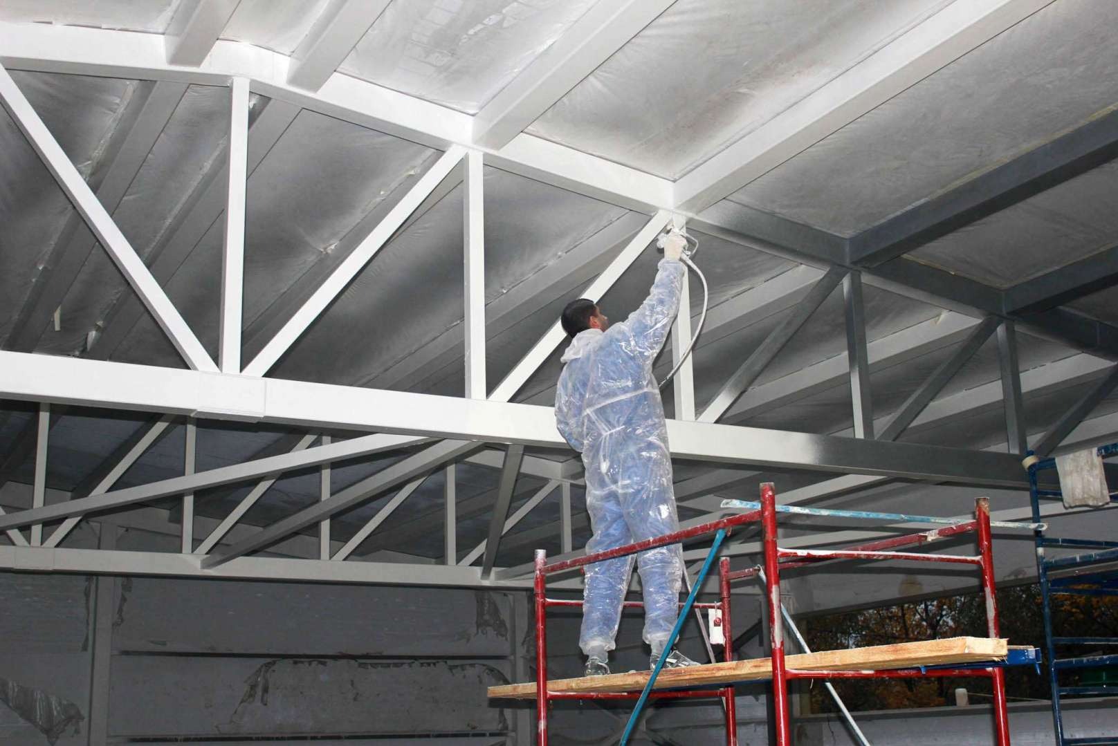 Pracownik w kombinezonie ochronnym maluje natryskowo konstrukcję dachu wewnątrz budynku, stojąc na rusztowaniu.