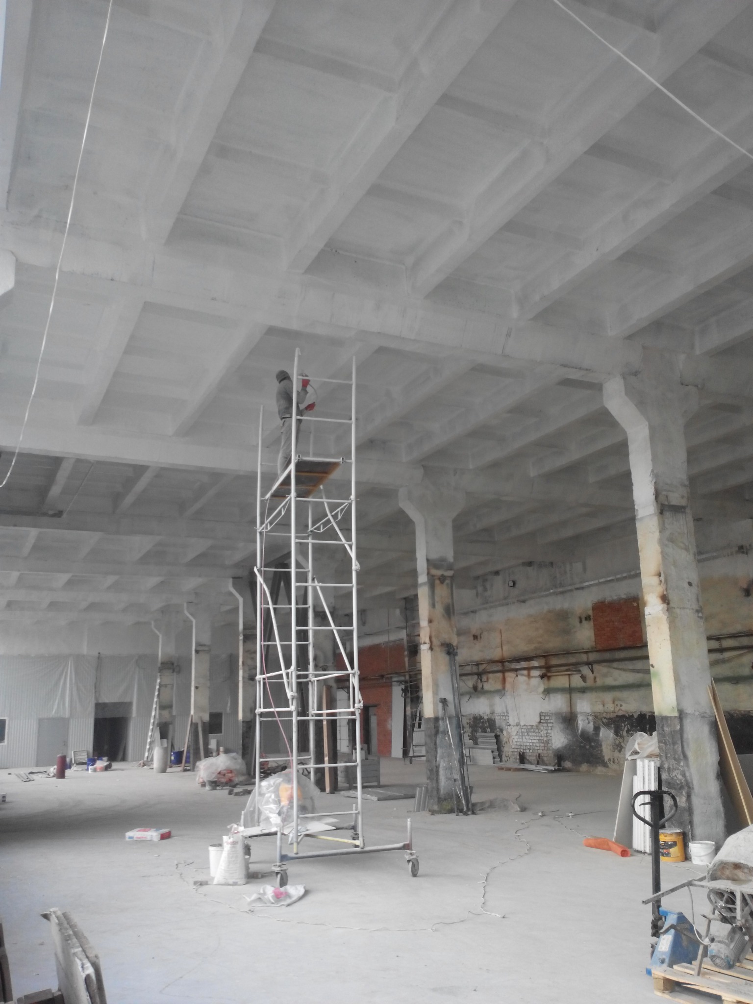 Pracownik na rusztowaniu maluje sufit w hali przemysłowej; widoczne betonowe filary i resztki starej farby na ścianach.