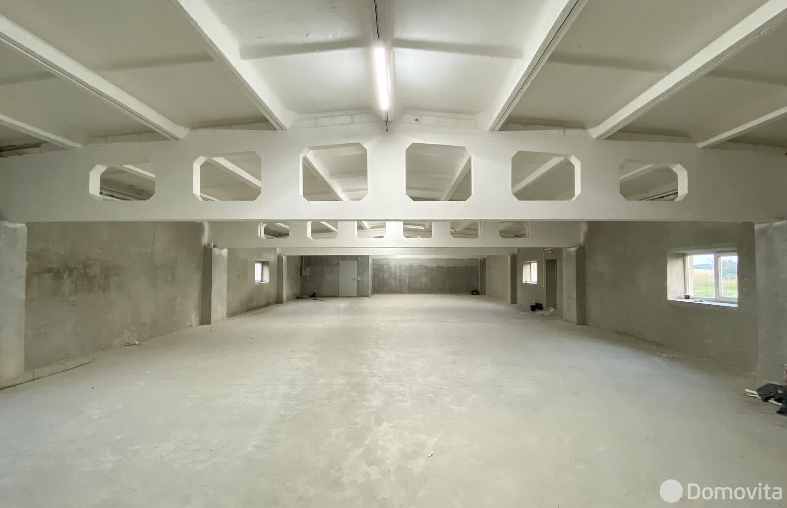 Wnętrze dużego, pustego pomieszczenia biurowego z surowymi ścianami, betonową podłogą, białymi belkami stropowymi i oświetleniem jarzeniowym, przygotowanego do malowania.