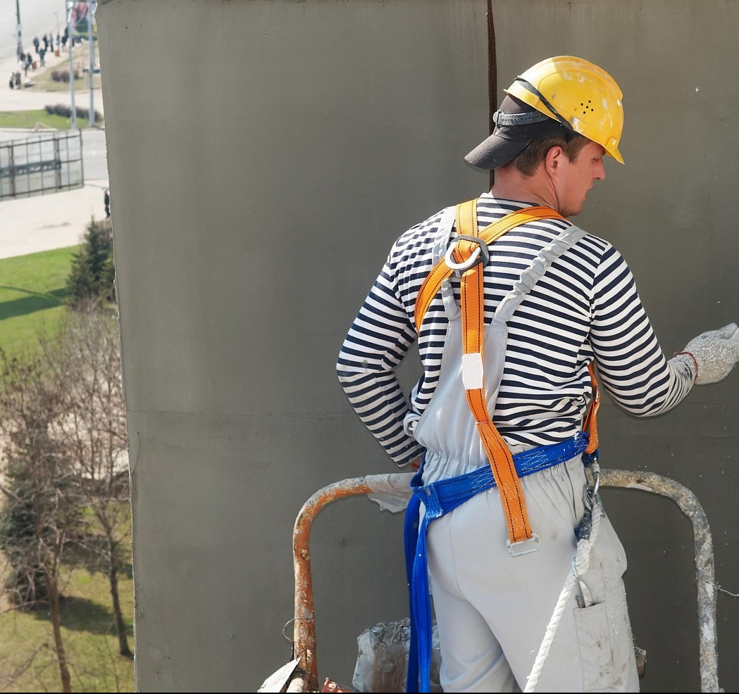 Pracownik w kasku i szelkach bezpieczeństwa, stojąc na rusztowaniu, nakłada szpachlę na szarą ścianę budynku. Widoczne drzewa i zabudowania w tle.
